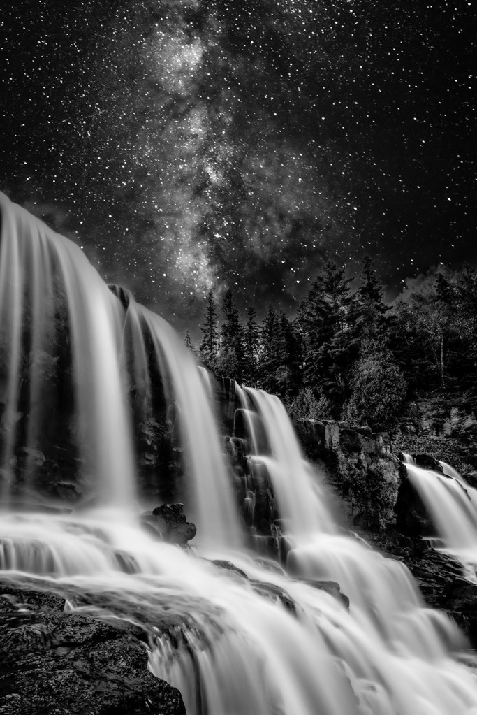 Black-and-white fine art photograph of a waterfall beneath the Milky Way. The long exposure captures the silky flow of water cascading over rocks under a luminous night sky, symbolizing the connection between Earth and the cosmos — where motion meets stillness and the Earth dreams the stars.